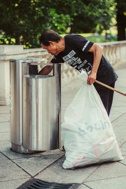 A man wearing a black T-shirt with a graphic design is disposing of waste into a stainless steel trash bin on a paved sidewalk. He is holding a large white trash bag in his left hand while leaning for