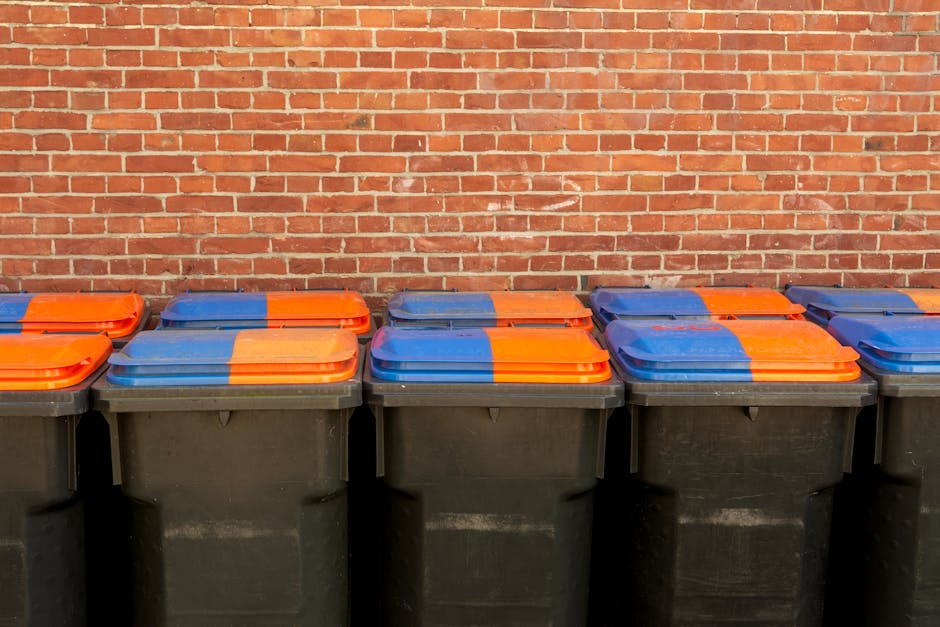 A man wearing a black T-shirt with a graphic design is disposing of waste into a stainless steel trash bin on a paved sidewalk. He is holding a large white trash bag in his left hand while leaning for