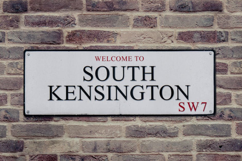 A street view of multi-story buildings in the Royal Borough of Kensington and Chelsea, featuring a mix of red brick and light-colored façades with ornate architectural details. The street appears cle