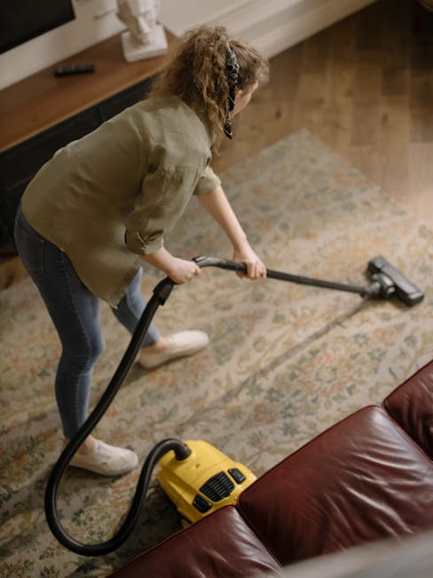 Close-up of a professional vacuum cleaner with a black and red body, transparent dust container filled with debris, and a flexible, patterned hose attached to the nozzle. The vacuum is positioned on a